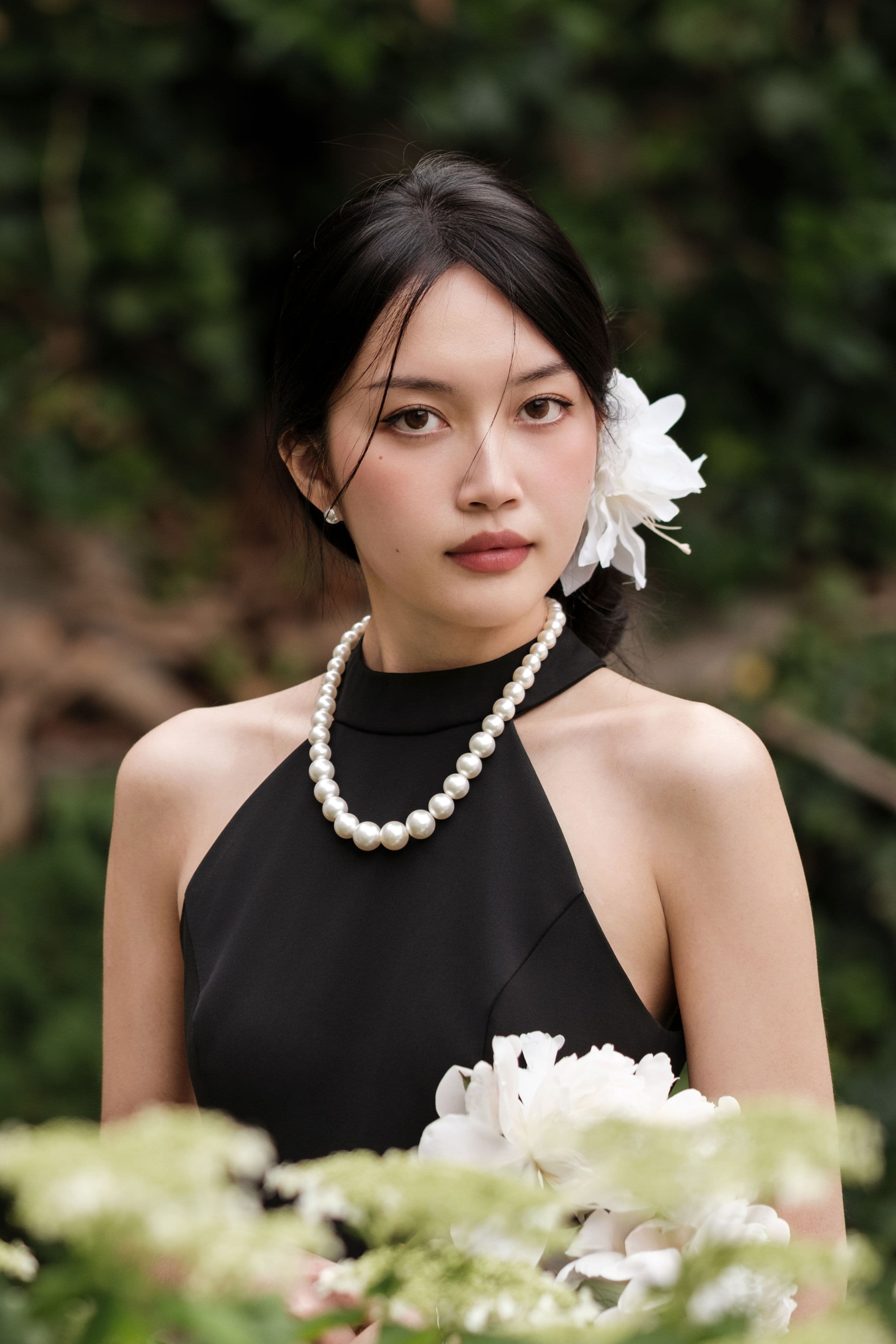 Portrait of a woman in a black halter dress with white flowers and a pearl necklace outdoors.