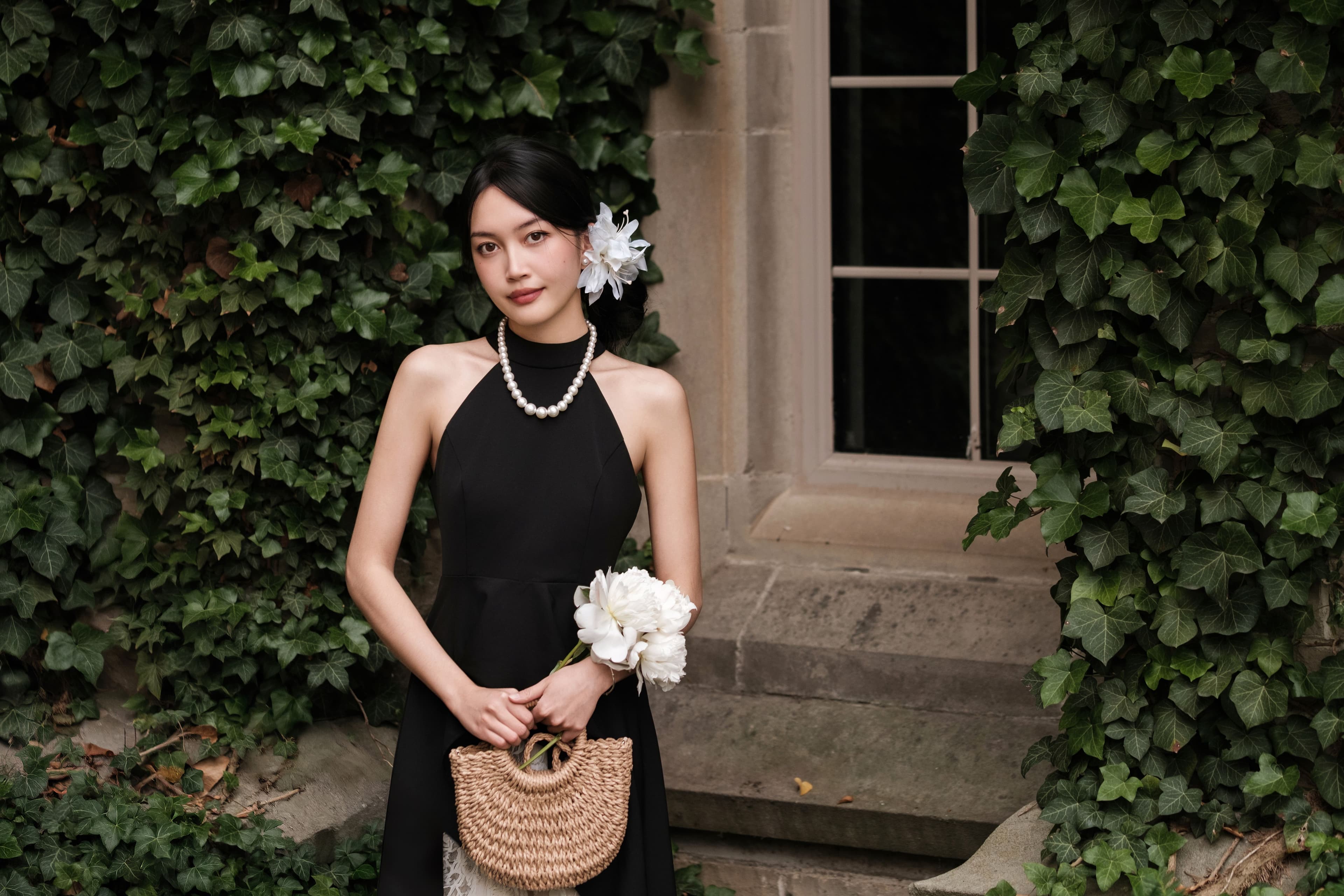 Side profile portrait of a woman wearing a black halter dress, pearl necklace, and a white lily, photographed against an ivy-covered wall at Western University, London, Ontario.