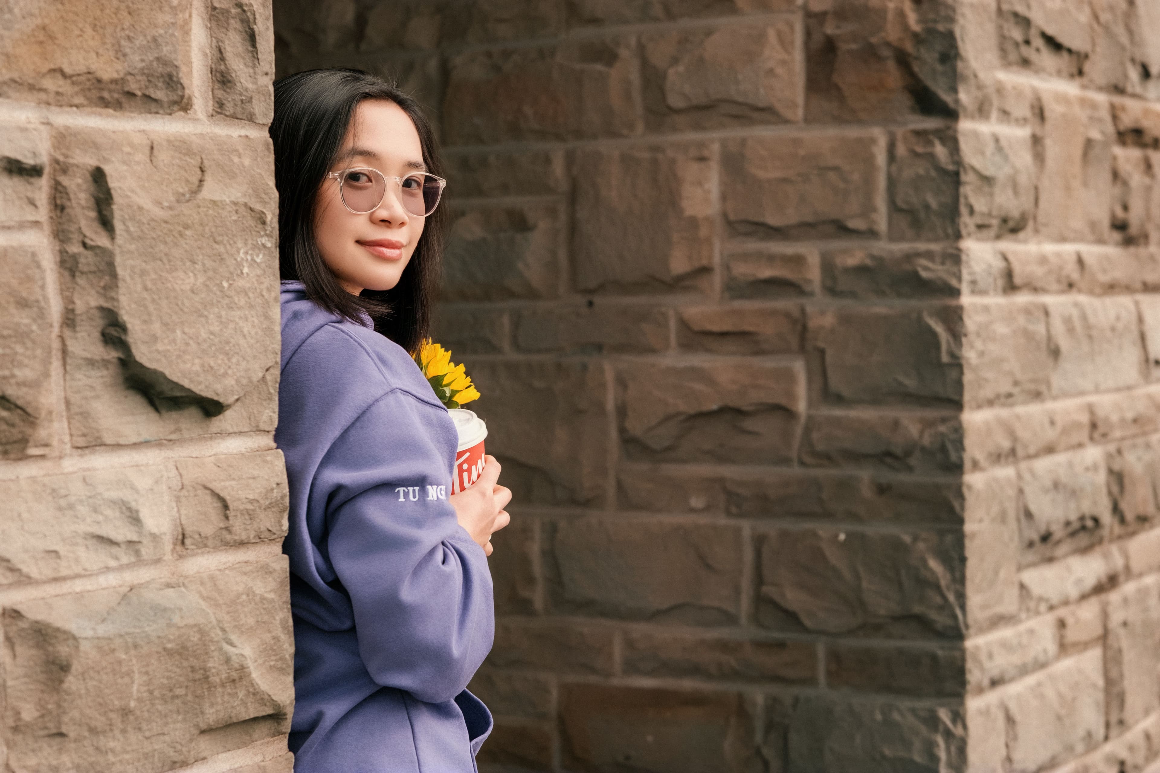 Graduate in a purple Western University hoodie leaning against a stone wall while holding a sunflower.