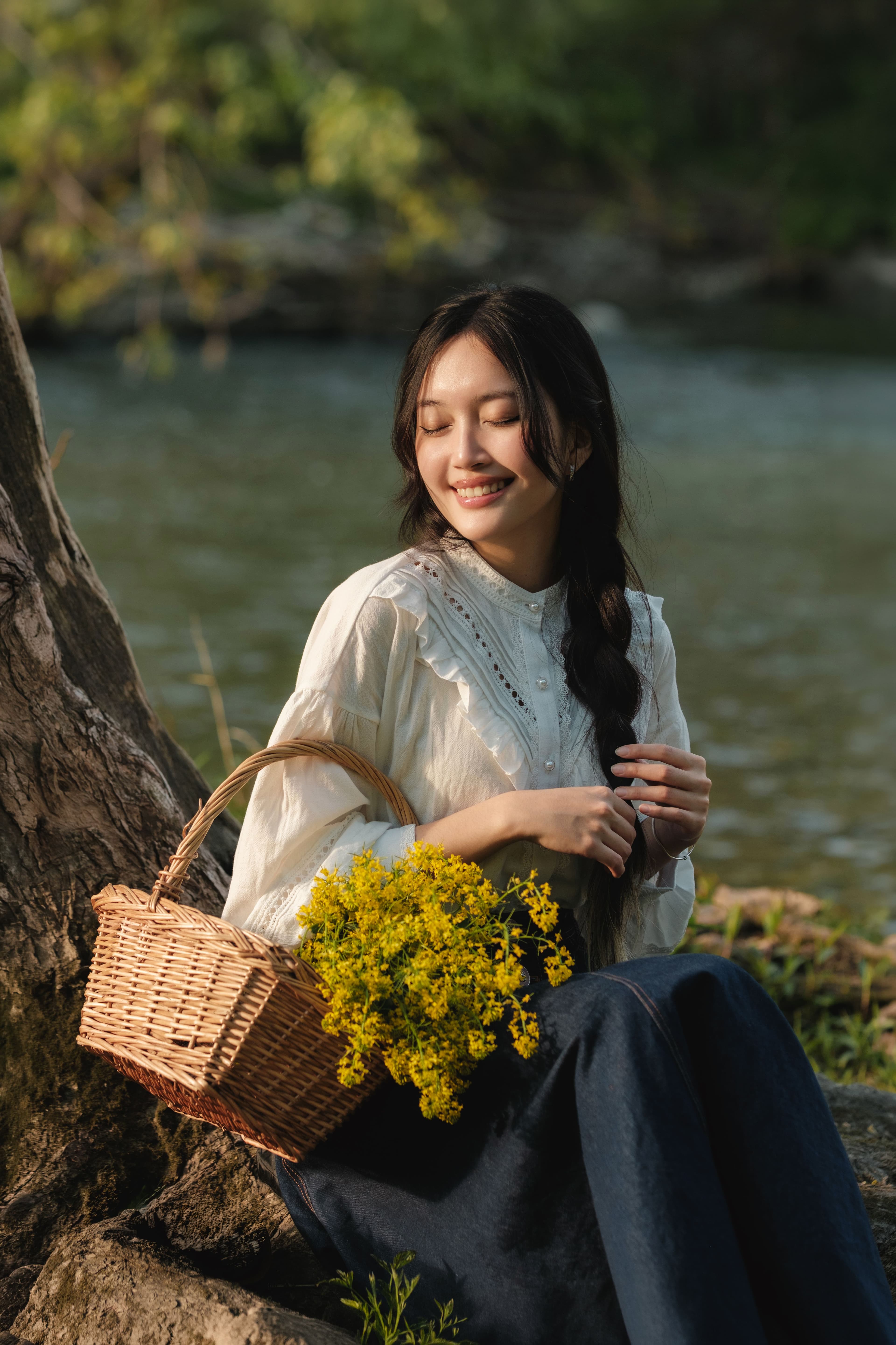 Natural light portrait of a smiling woman sitting beside the Thames River with a basket of yellow wildflowers at Ross Park, London, Ontario, during golden hour.